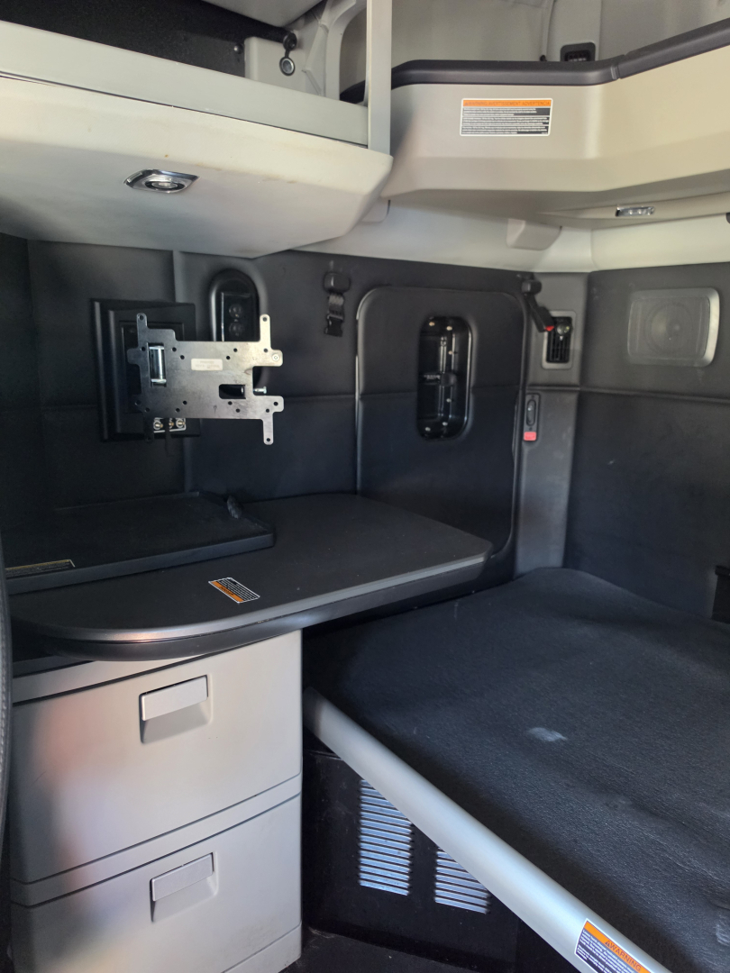 Cab interior of a 2023 Kenworth T680 sleeper showing a mounted TV bracket and storage drawers in the bunk area with a black seat/bed surface.