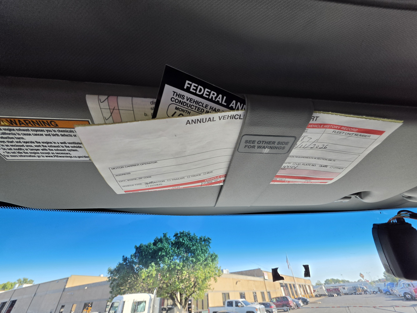 Cab interior roof area with sun visors and paperwork tags; visible label text includes warnings and federal compliance cards.