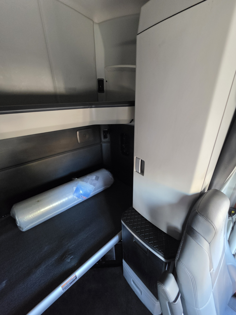Cab interior of a Kenworth truck sleeper with a small shelf and storage area, a rolled-up item lying on a black surface, and a light gray cabinet near the driver seat.