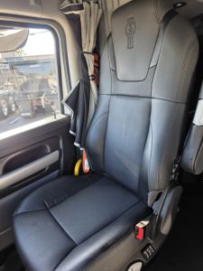 Vehicle cab seat in a 2023 Kenworth T680 truck, driver seat with embroidered Kenworth logo, gray upholstery, window and curtain visible.