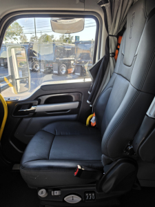 Truck cab seat interior showing driver seat and door panel of a Kenworth T680 conventional sleeper, with window view of other trucks outside.