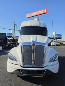 Kenworth T680 front view showing large chrome grille, headlights, and Kenworth dealership sign in background.
