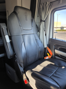 Kenworth T680 truck interior seat and door area with black leather seat and seatbelt, driver’s window, curtain in sleeper area, gray dashboard edge visible.