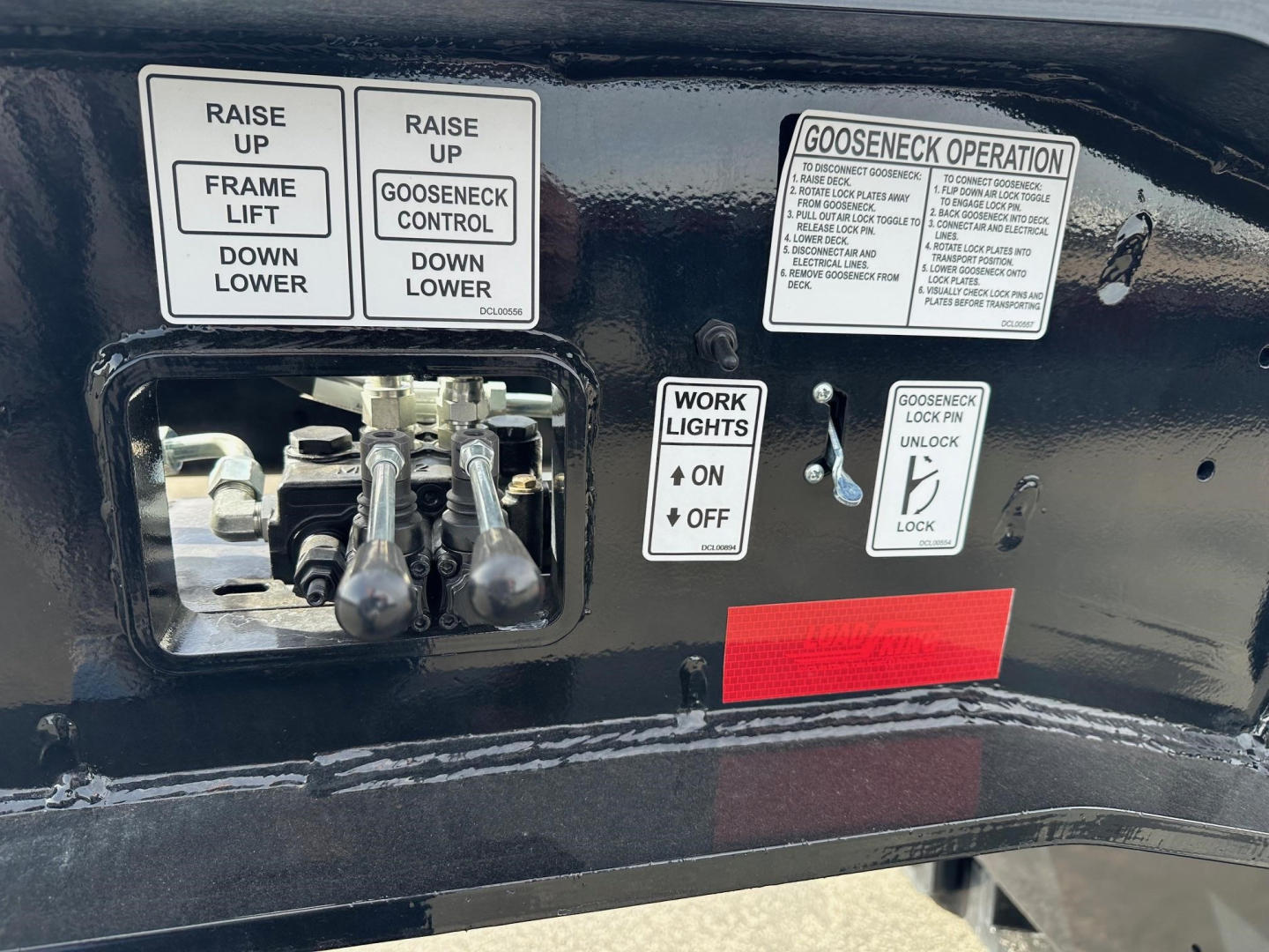 Control panel on a Load King 55 Ton lowboy trailer showing levers and stickers. Visible text includes phrases: 'RAISE UP', 'FRAME LIFT', 'GOOSENECK CONTROL', 'WORK LIGHTS ON OFF', 'GOOSENECK OPERATION' and a long instructional label. The VI