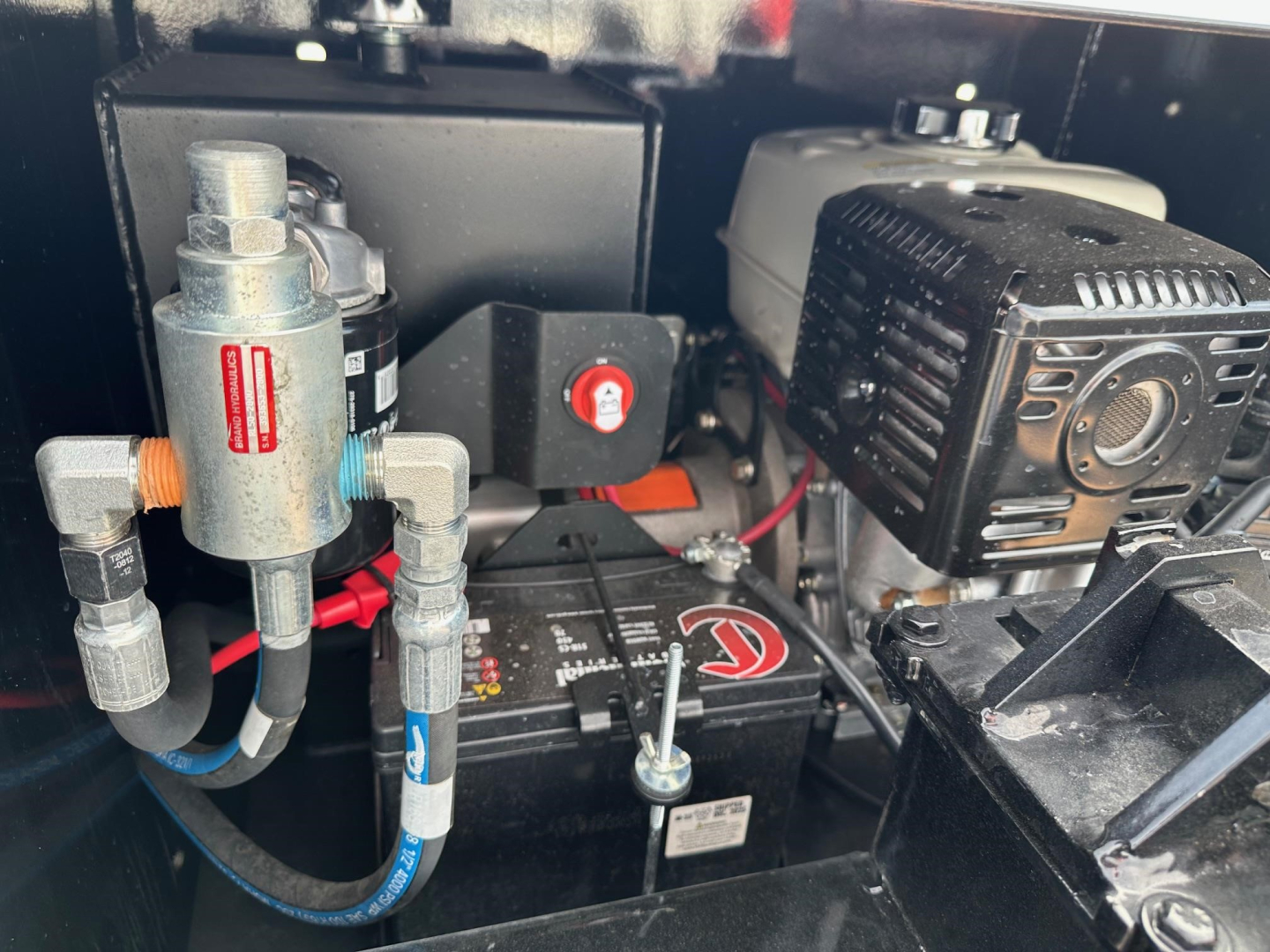 Lowboy trailer engine bay showing fuel filter, battery, hydraulic tank, and small gasoline engine with starter. Visible labels and fittings, compact layout.