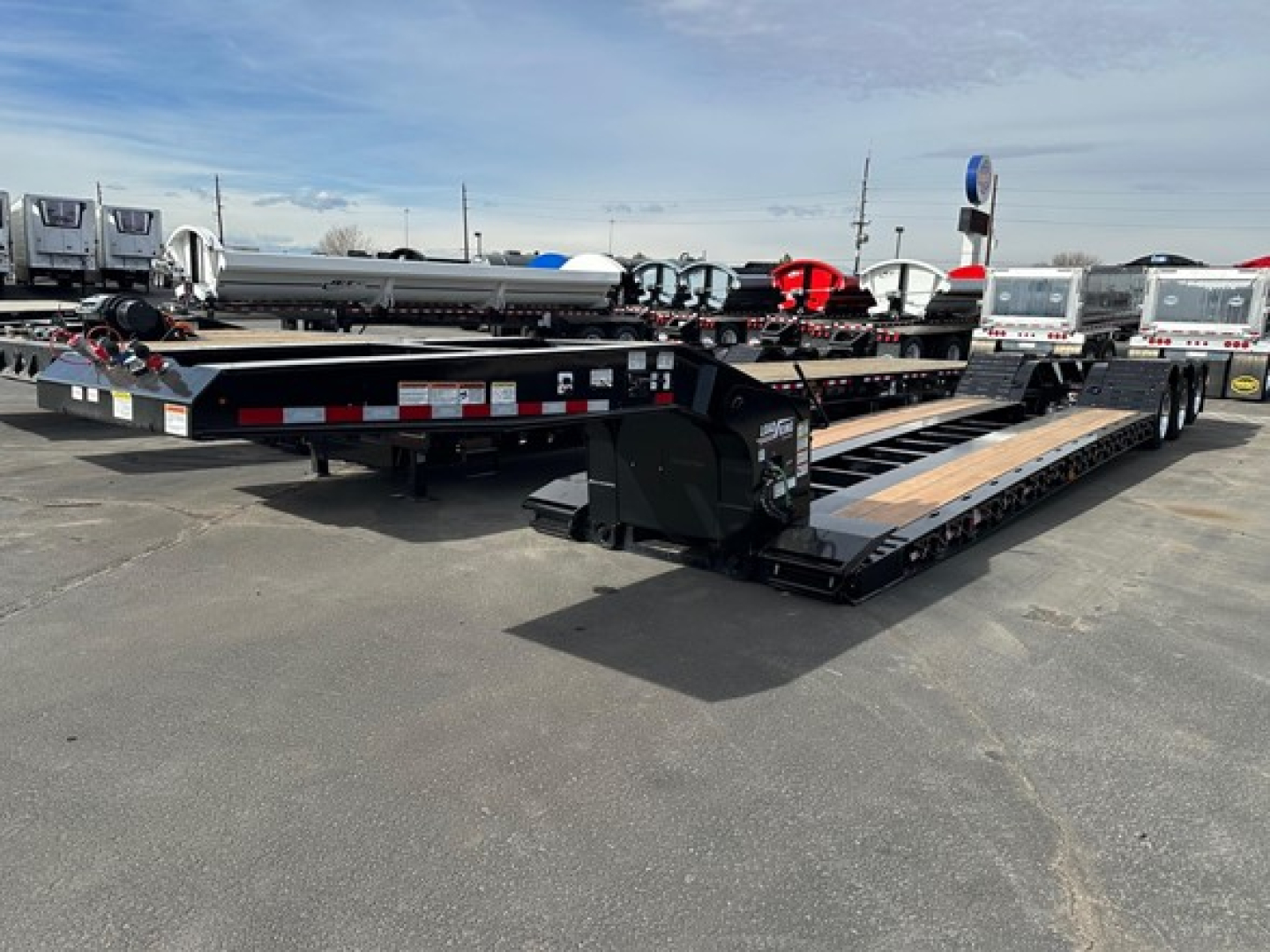 Lowboy trailer in a dealership lot showing black steel deck with wooden planks, hydraulic ramps, and stickered markings along the sides.