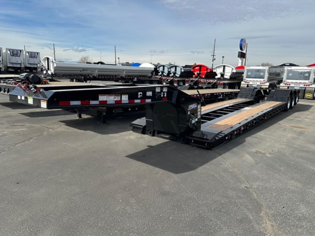 Lowboy trailer in a dealership lot showing black steel deck with wooden planks, hydraulic ramps, and stickered markings along the sides.