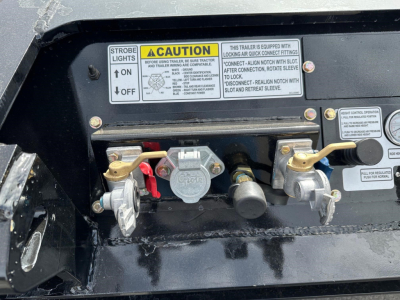Trailer components showing hitch block and warning labels; strobe light switch label, caution label, two silver hitch latches, and hydraulic coupler with red handle; warning stickers and blue-on-black serial text.