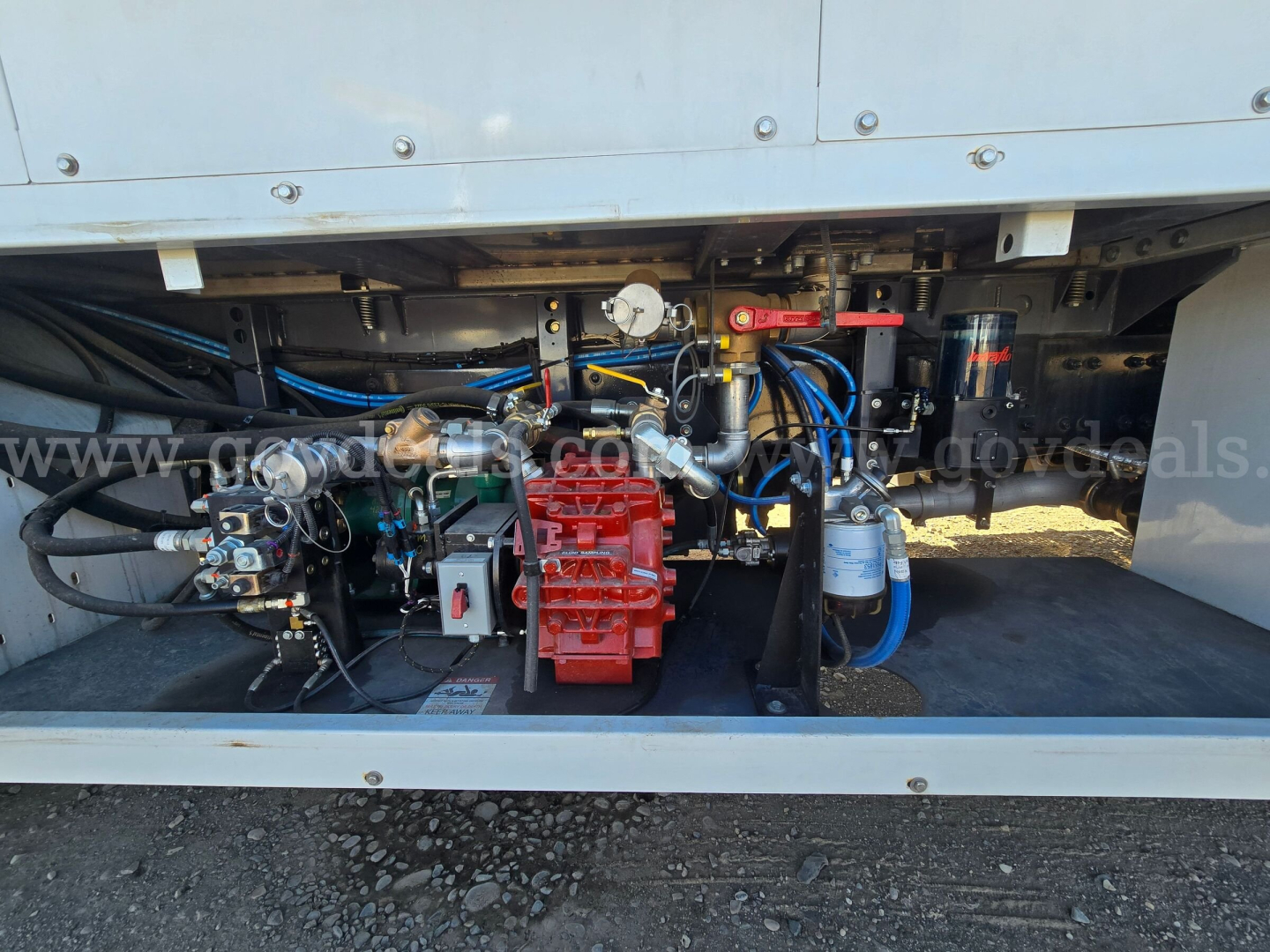 Truck engine bay showing auxiliary engine and components under freight trailer with red hydraulic pump, blue hoses, and gauges.