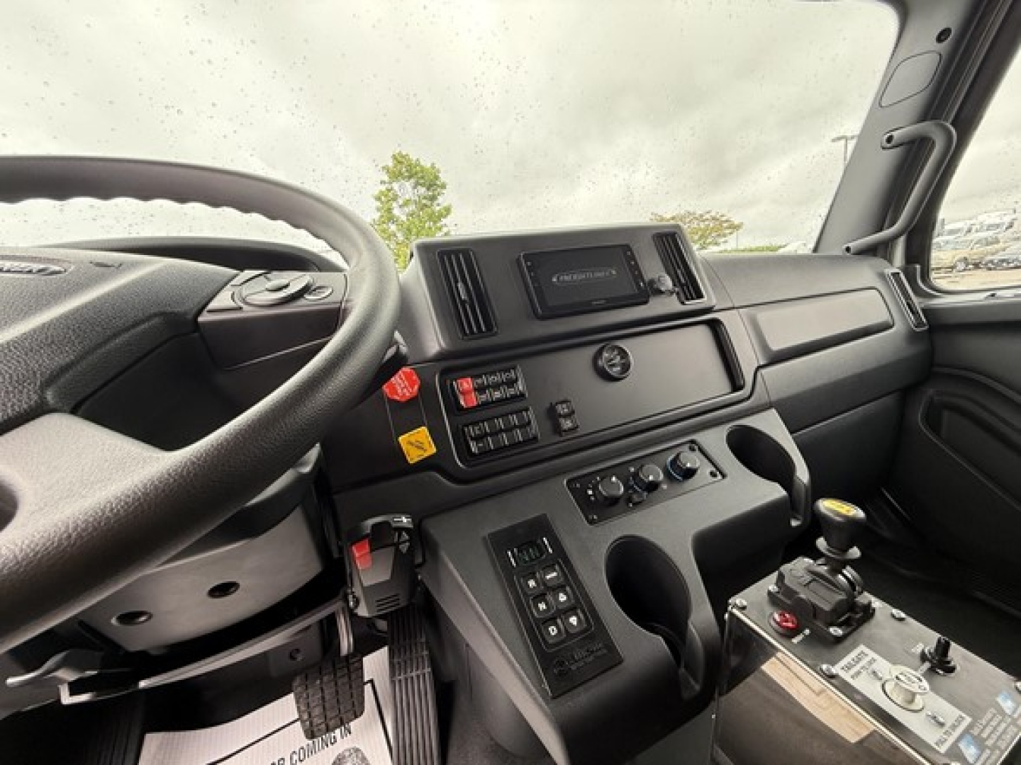 Truck cab dashboard interior showing steering wheel, digital screen, control panel, and joystick gear selector.