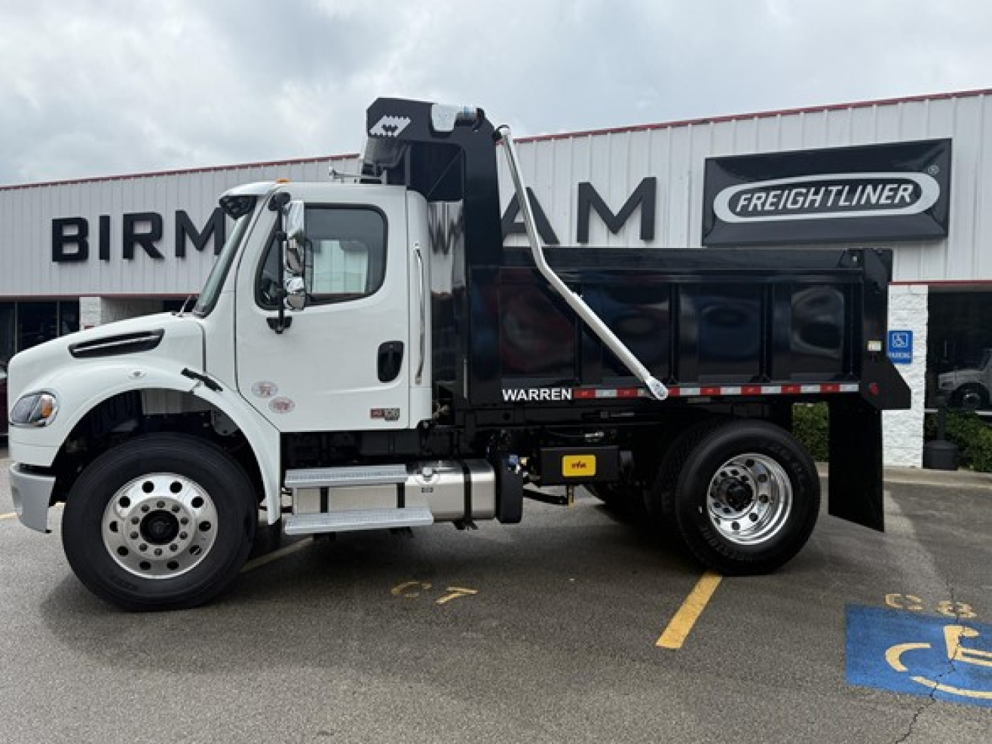 Dump truck side view showing Freightliner Business Class M2 106 with dumping bed, white cab, black dump body, and Warren hoist. Visual text includes Freightliner logo on building, stock number 327489