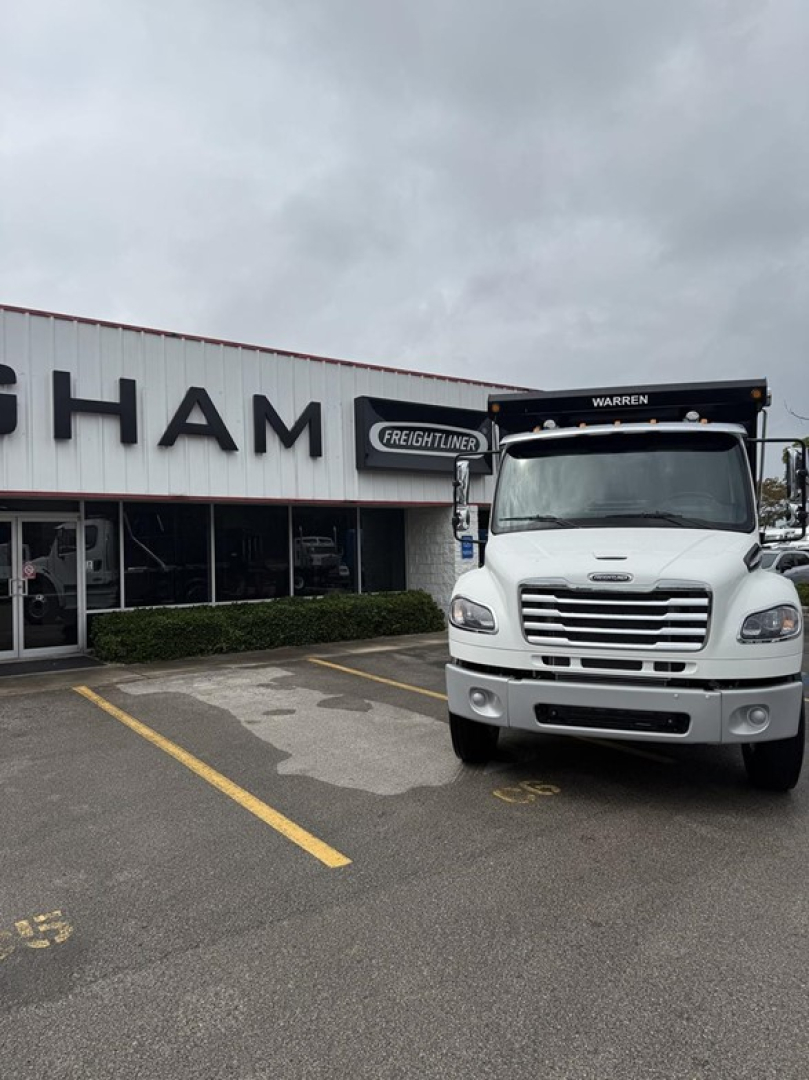  Freightliner Business Class M2 106 dump truck in front of dealership, front view visible with grille and dump bed.