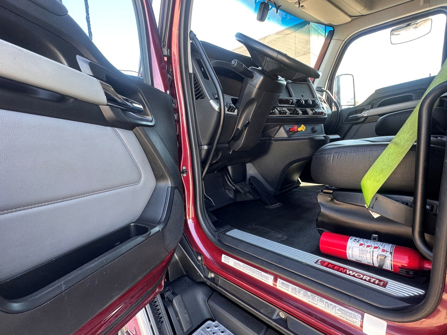 Interior of a Kenworth T680 truck cab with driver seat, steering wheel, dashboard, and red exterior door sill. Visible label areas and a fire extinguisher mounted near the door.