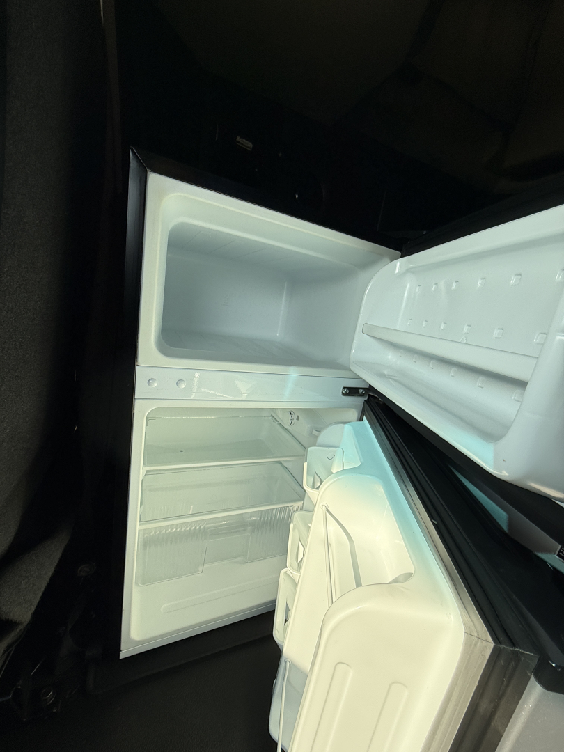 Refrigerator inside a Kenworth T680 cab with open doors showing empty interior compartments and shelves.