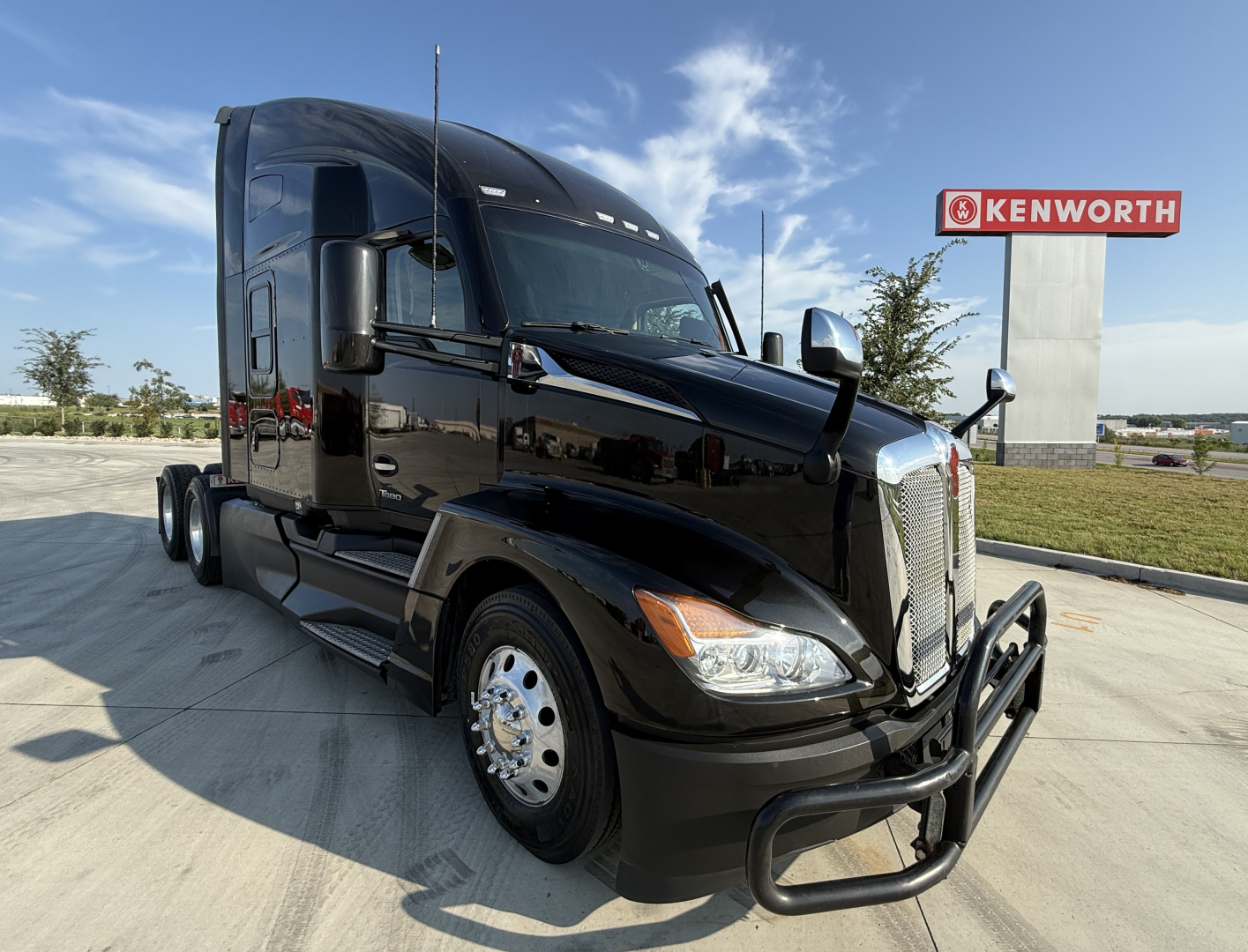 Kenworth T680 tractor-trailer in black exterior viewed from front-left, with chrome bumper guard and Kenworth dealership sign in background.