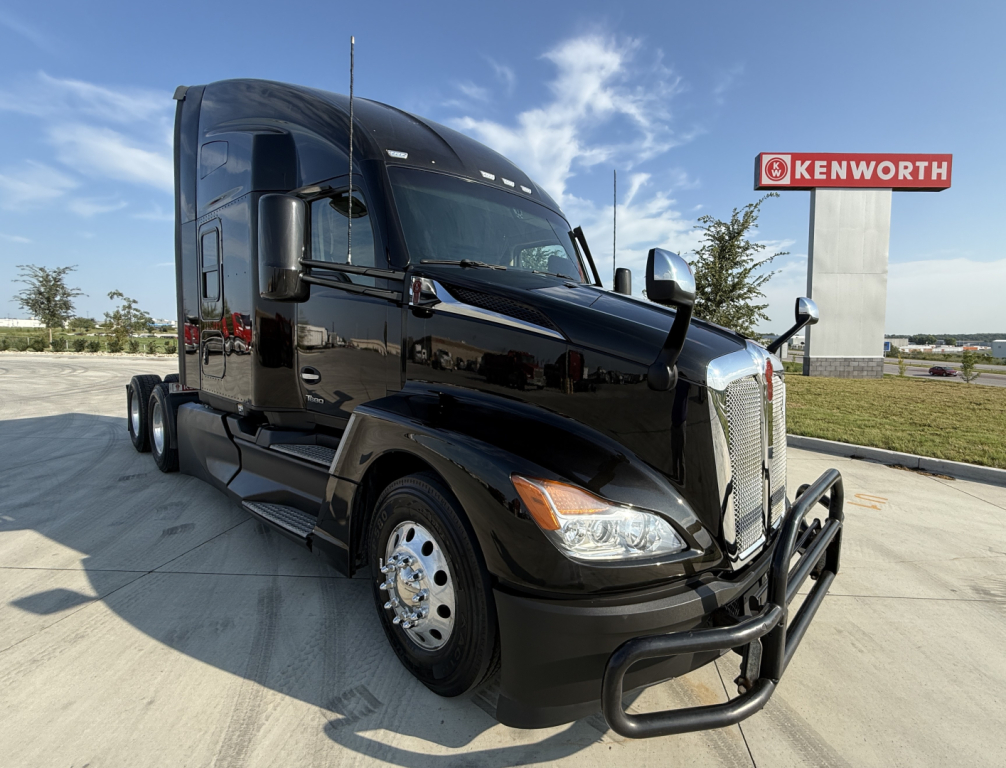 Kenworth T680 tractor-trailer in black exterior viewed from front-left, with chrome bumper guard and Kenworth dealership sign in background.