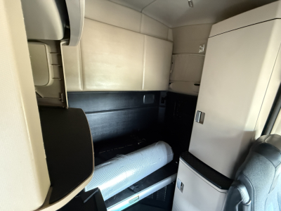 Kenworth T680 cabin interior showing sleeper area and storage compartments with light beige upholstery and a white refrigerator.