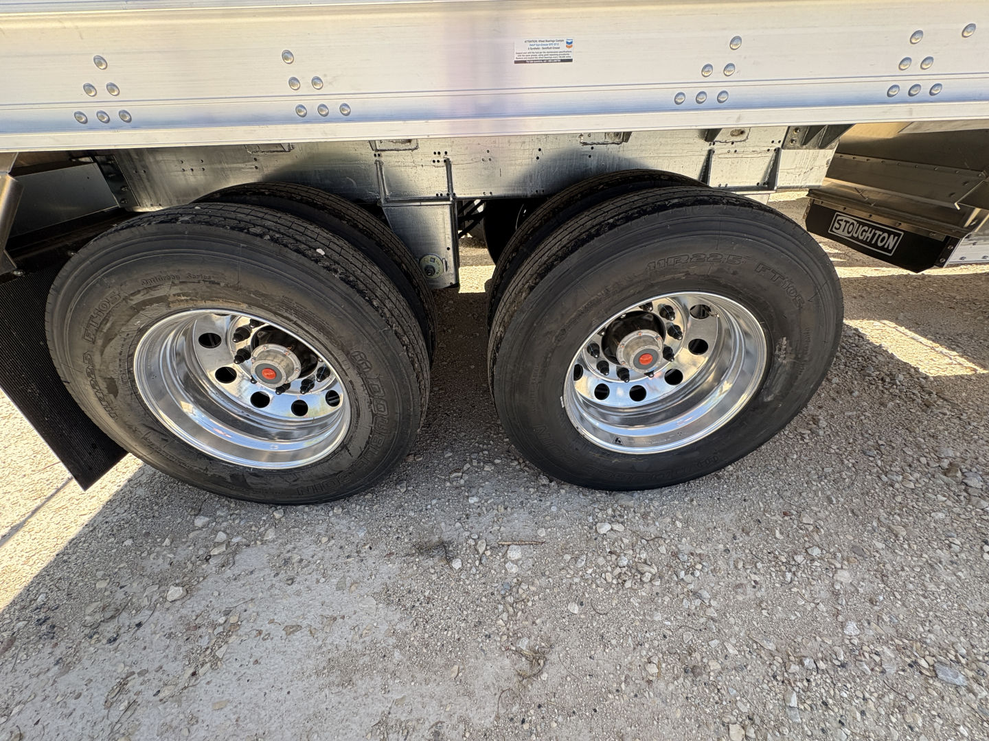 Twin axle trailer wheels with Stoughton logo visible on the frame; sidewall riveted aluminum with 74" corrugated sides; part of a 42' aluminum grain hopper trailer.