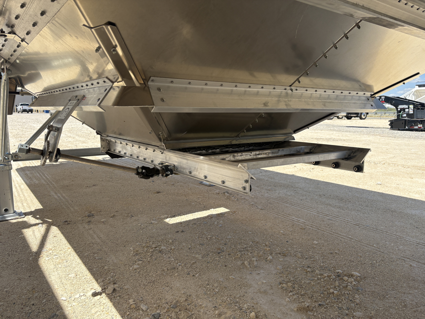Alum grain hopper trailer undercarriage with corrugated sidewalls and discharge chute visible; metal framework, bolts, and winch linkage near the underside.