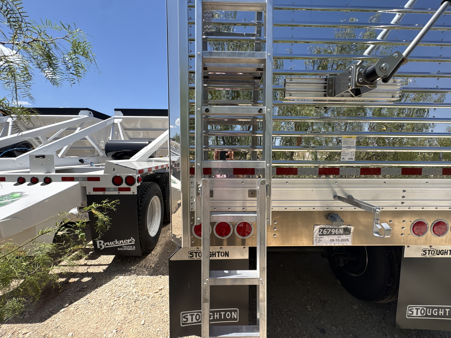 Rear view of a Stoughton grain hopper trailer with a vertical ladder, corrugated sidewalls, and taillights; visible are Stoughton branding, license plate area, and reflective markers.