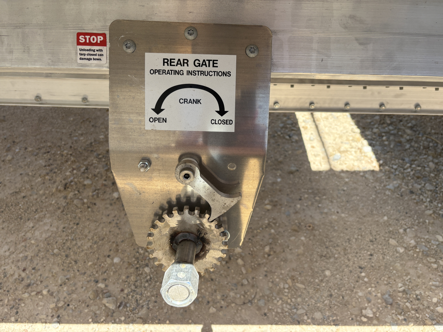 Trailer rear gate crank with operating instructions plate and gear handle visible on aluminum grain hopper trailer.