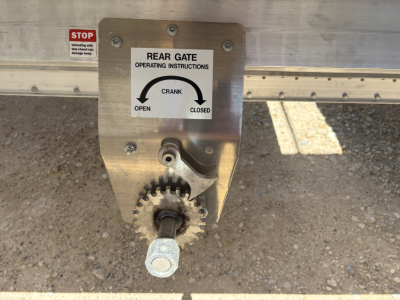 Trailer rear gate crank with operating instructions plate and gear handle visible on aluminum grain hopper trailer.