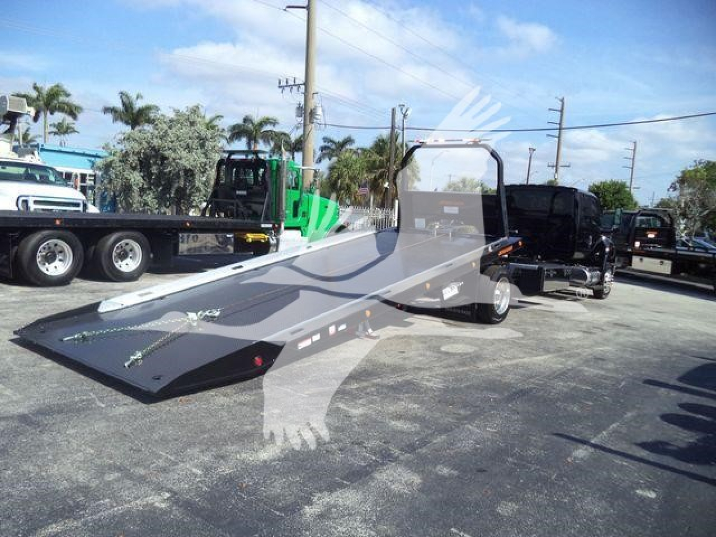 Flatbed tow truck chassis with ramp in foreground and other trucks in lot; MVEXCABBLKSHRKFM stock number visible.