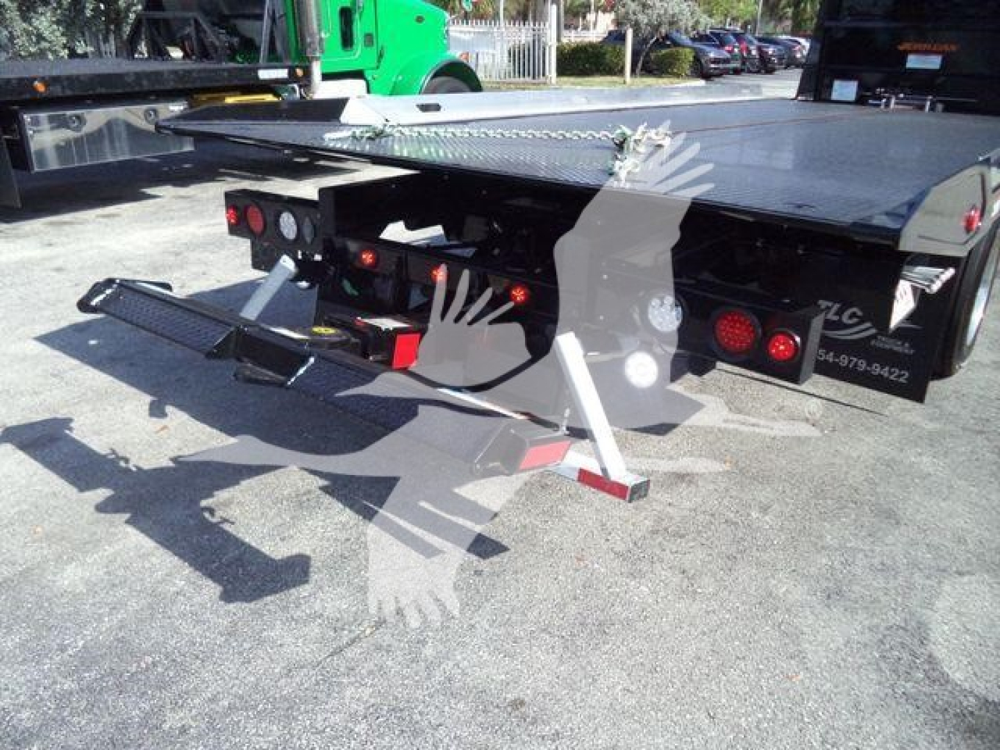 Truck flatbed with rear access steps and rollback equipment visible, truck tail lights and reflective markers on the bumper, hydraulic components, and a black flatbed surface.