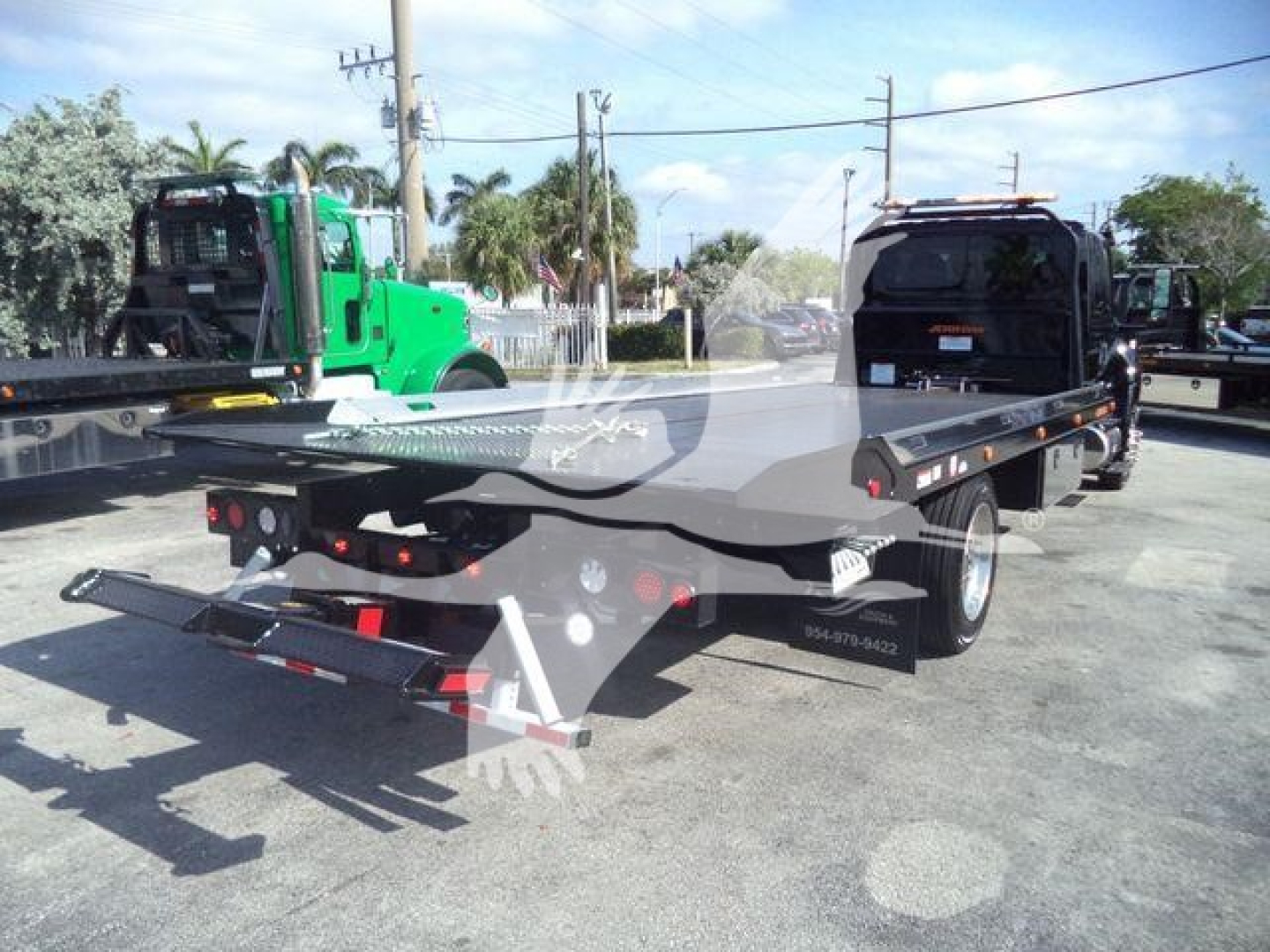Flatbed tow truck in a dealership lot with green equipment in background and clear sky.
