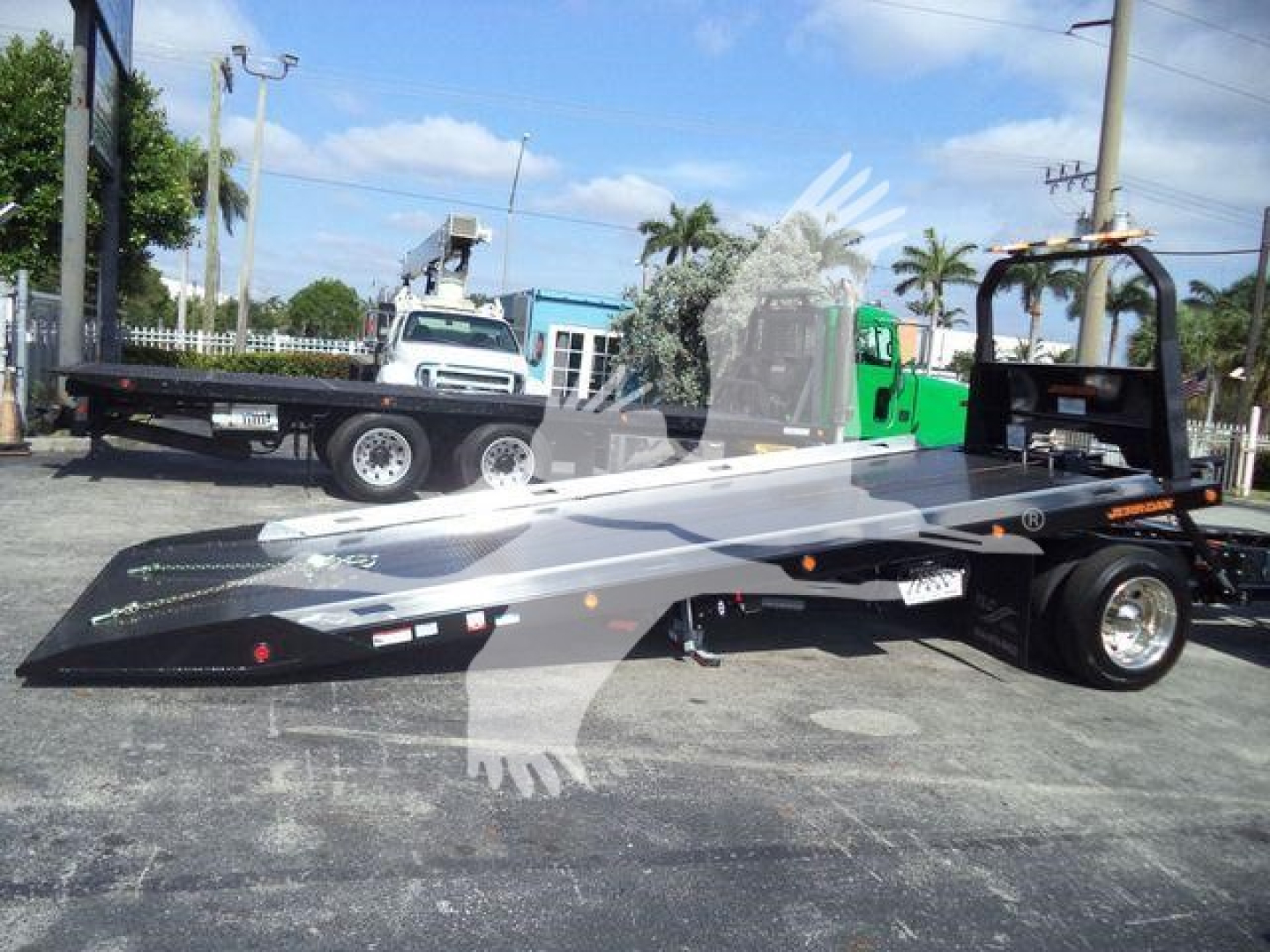 Inline tow truck trailer with a folded-down flatbed in a dealership lot. The deck has various reflective decals and warning labels. A white cab and green equipment are in the background.