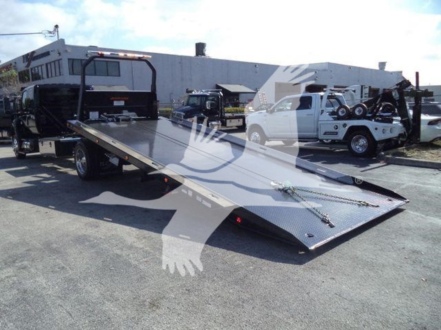 Flatbed tow truck trailer viewed from side with ramp down, black steel deck, white International MV pickup truck in background, rolling equipment and trucks nearby.