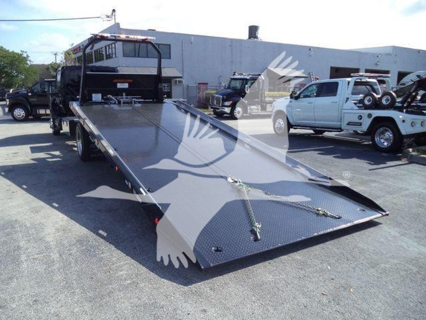 Flatbed tow truck trailer in dealership lot with other trucks in background and a watermark across the image.