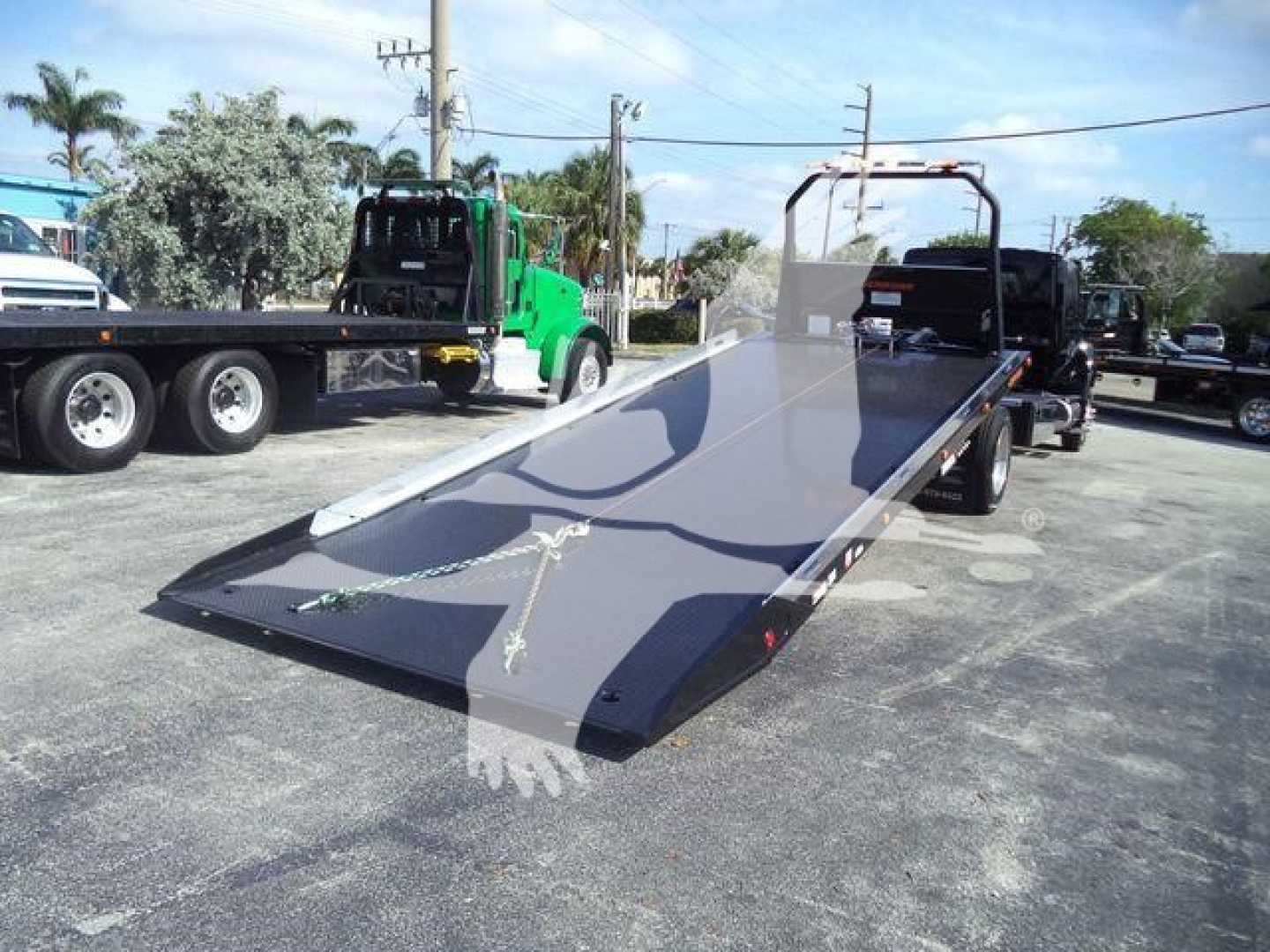 Flatbed tow truck trailer ramp with black surface and metal edge in a lot; trucks and equipment in background.