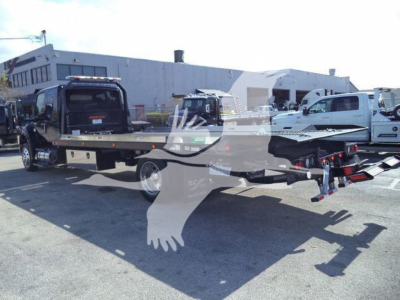Flatbed rollback truck with a black tow truck on the bed, white service vehicles and a large industrial building in the background.