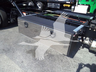 Aluminum toolbox mounted on the chassis of a heavy truck, showing a rectangular metal storage box with two circular lock handles and mounting hardware on a truck frame.