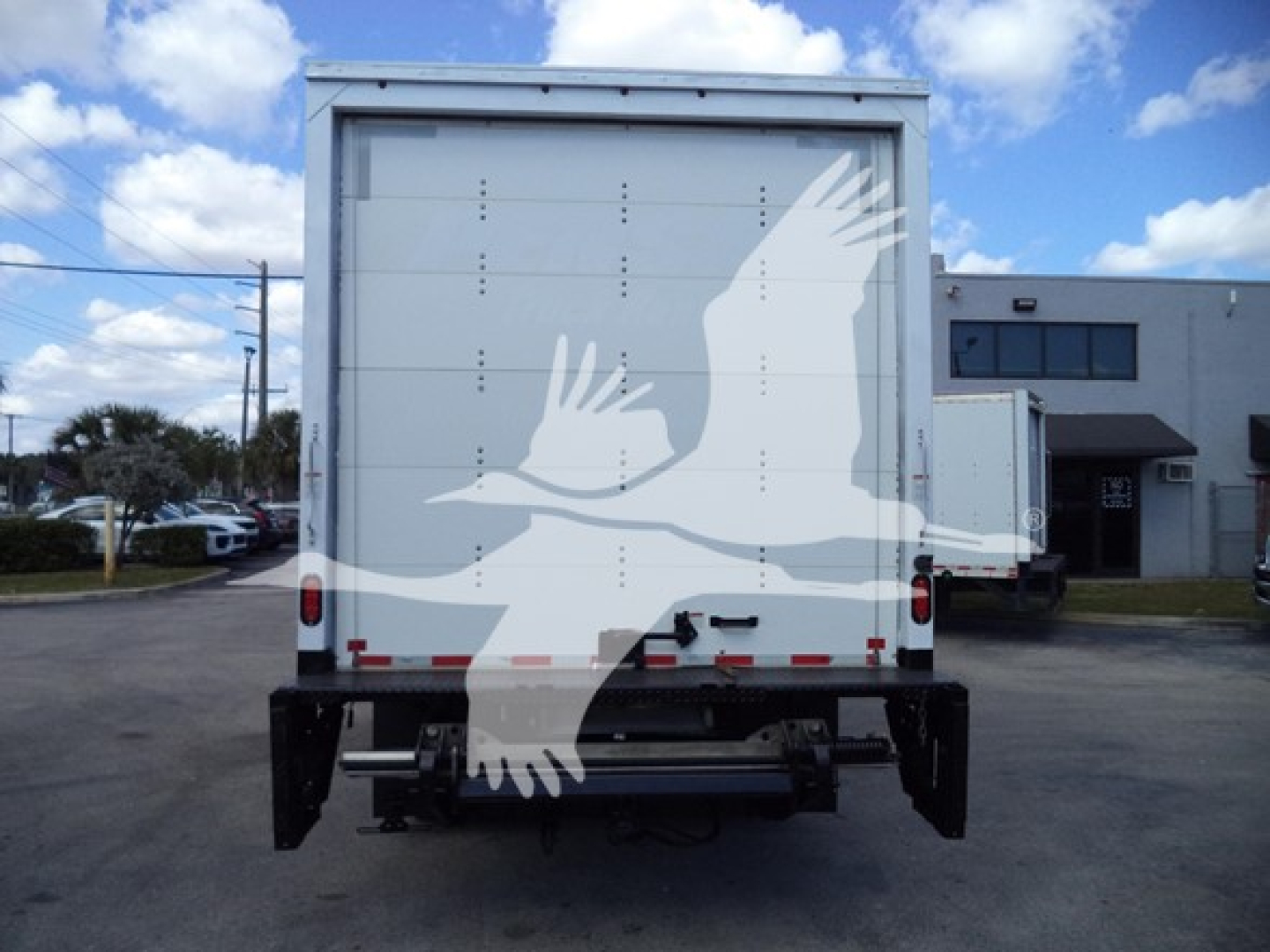 Rear view of a box truck with Isuzu NPR-HD cargo area; visible are the back doors, bumper, and undercarriage; clear sky and a building in the background.