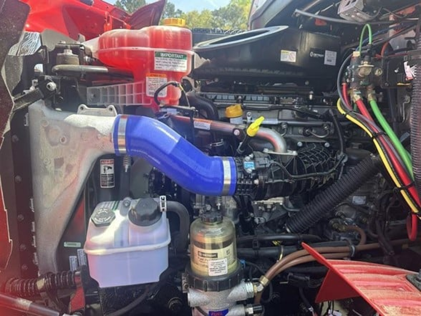 Truck engine bay showing blue intercooler pipe, coolant reservoir, yellow dipstick, and various hoses and wiring in a Western Star 47X engine compartment.
