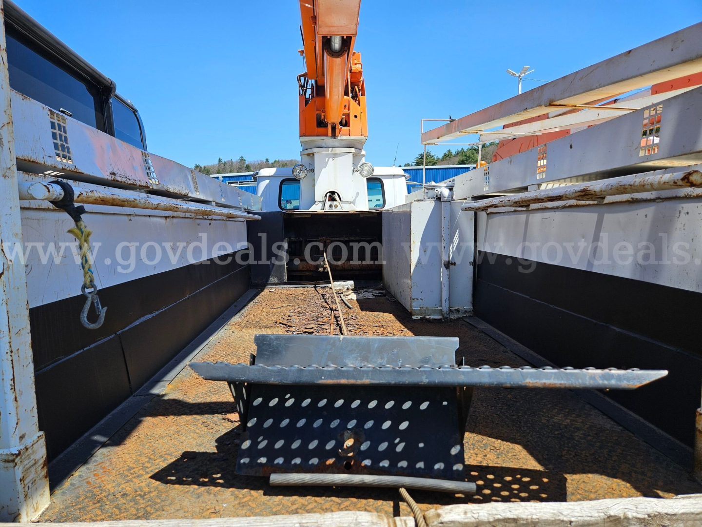 Truck bed view of a 1995 International 4900 boom truck, showing metal bed, perforated step piece, rusted deck plates, and large orange boom in the background.