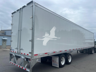 Trailer side view showing a silver Vanguard Maxcube trailer with plain sides, rear double doors, dual rear axles, and a white bird logo.
