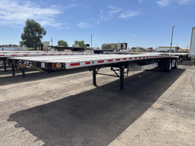 Flatbed trailer low-angle view showing long metallic platform, undercarriage legs, and wheel axles with dual rear tires.