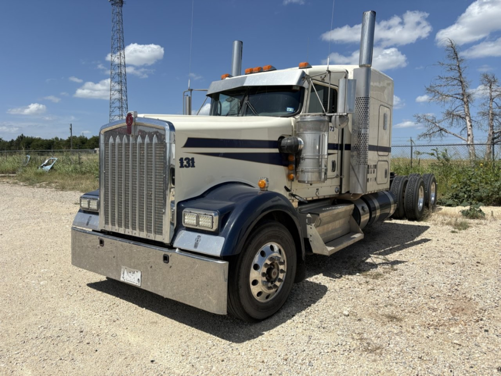 Kenworth conventional sleeper truck in gravel lot showing front grille, headlights, chrome bumper, dual exhaust stacks, and blue/gray paint with 131 decal.