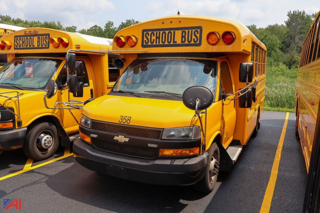 Front view of a yellow Chevrolet Thomas Express mid-size school bus with 'SCHOOL BUS' sign, 358 number on hood, mirrors, and other yellow buses in background.