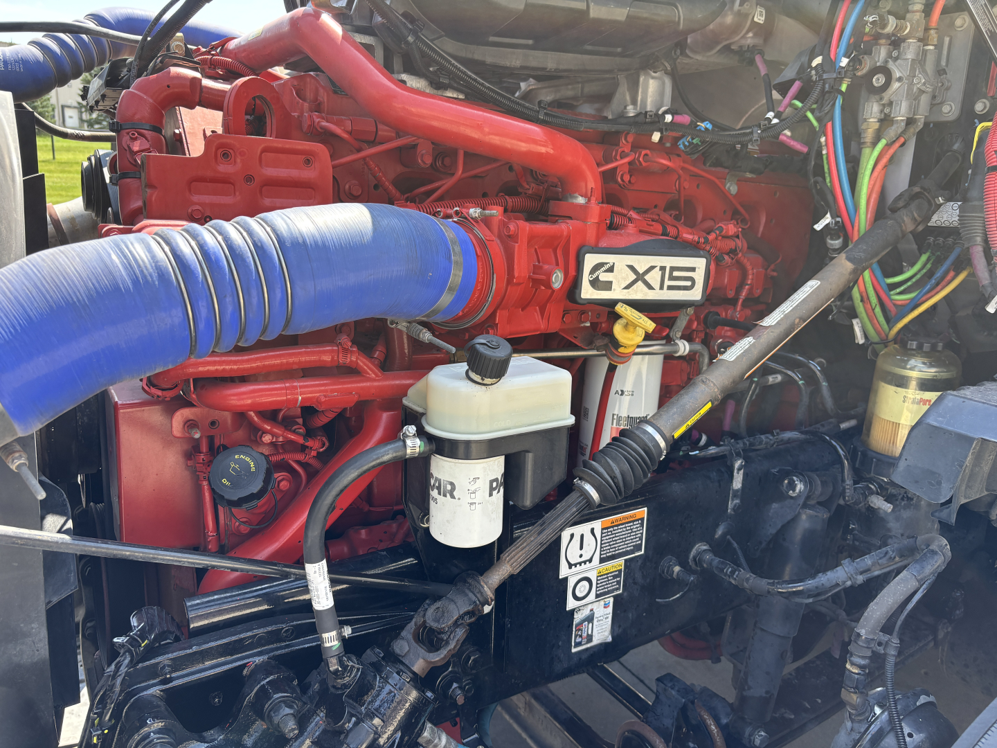Kenworth T680 engine bay showing X15 badge and red engine with blue intake hose and various wiring and fluid reservoirs.