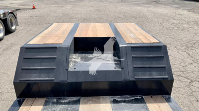 Lowboy trailer bed with wooden deck and black steel sides, central cutout with glass window and side vents, branding not visible.