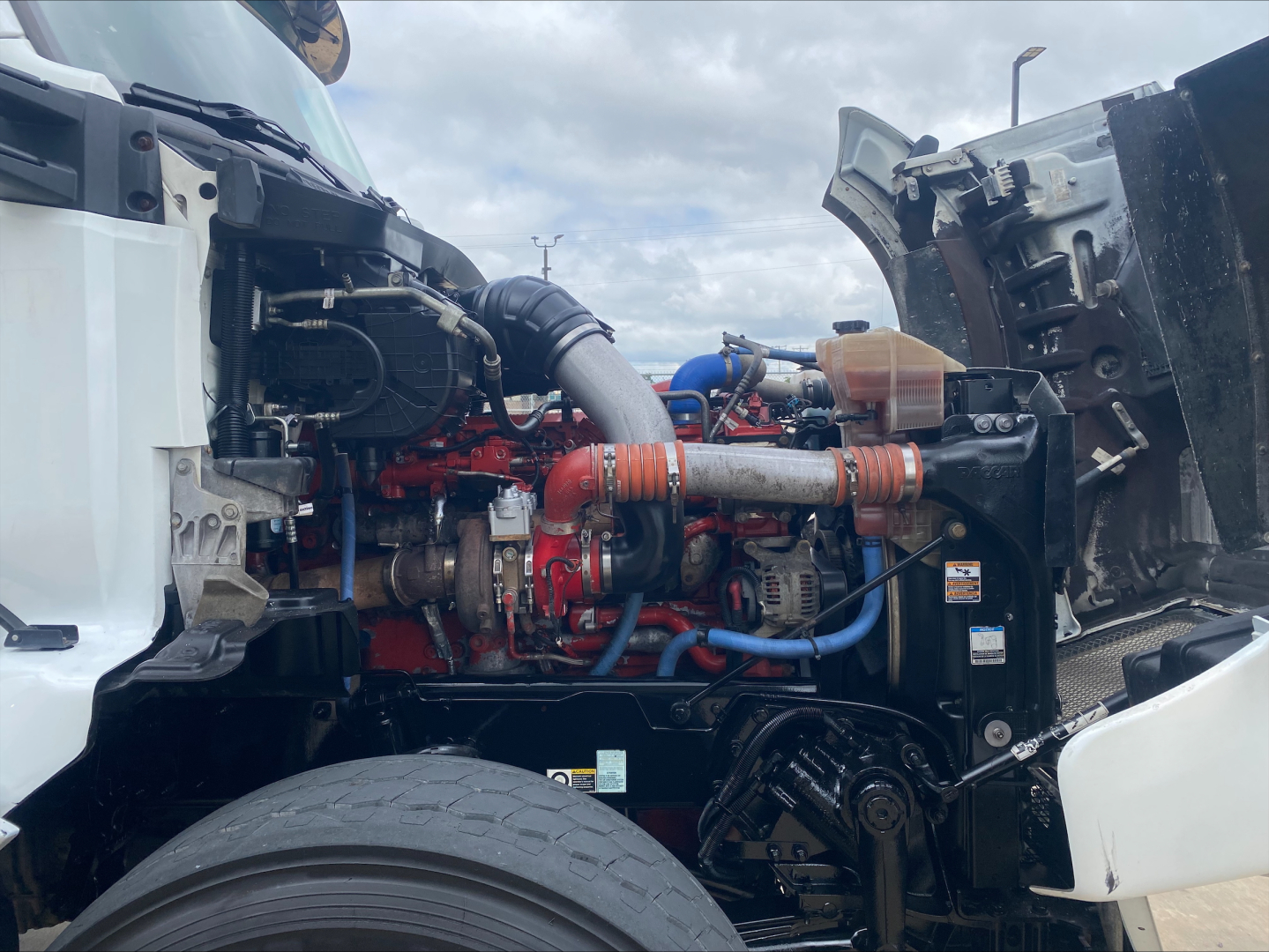 Kenworth T880 engine bay showing diesel engine with red components, blue hoses, and air intake; visible front wheel and open hood area.