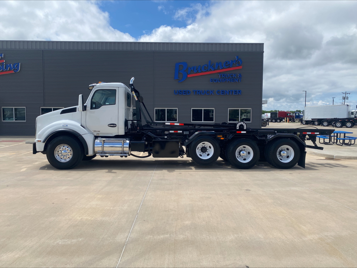 Kenworth T880 rolloff truck side view showing chassis and dual rear axles with flatbed; white cab with chrome fuel tanks.