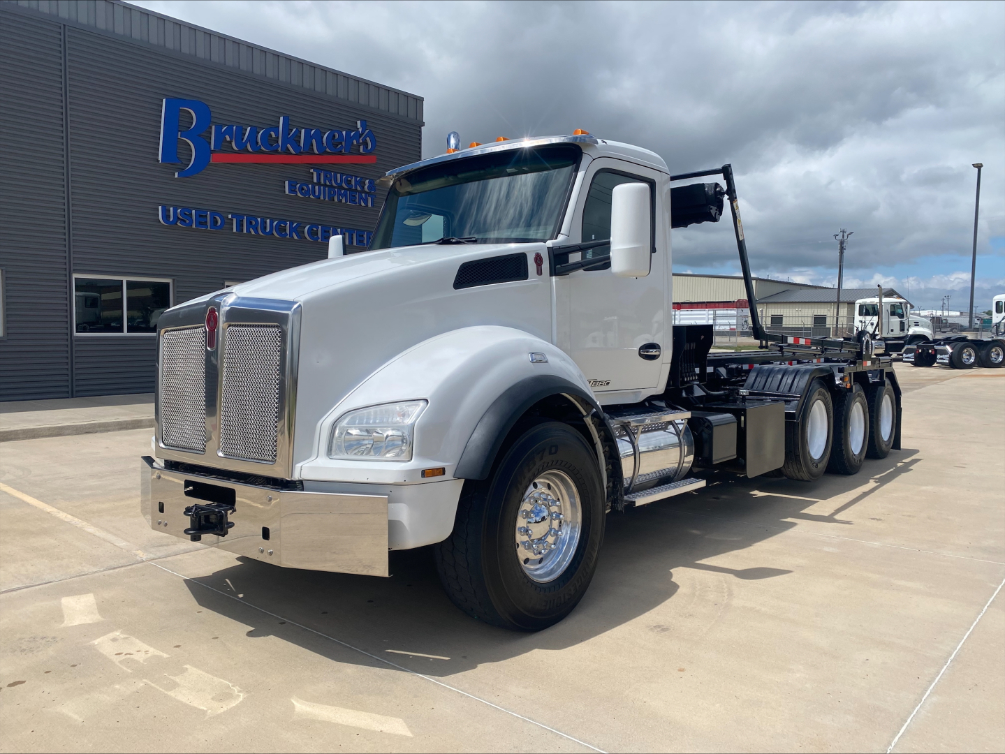 Truck cab of a 2018 Kenworth T880 with a heavy-duty chassis, front bumper, grille, and wheel wells visible; text on building reads 'Truck' and 'Equipment' but no legible model details on the vehicle from this angle.