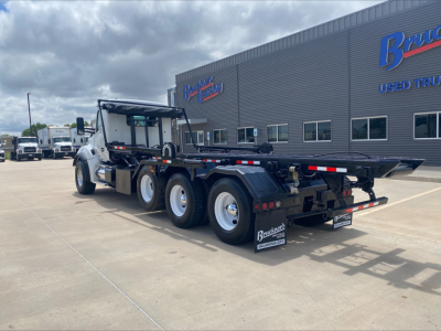 2018 Kenworth T880 roll-off truck with flatbed, long wheelbase, dual rear bogies, rear view showing rear bumper and mud flaps. visible text on building reads 'Bra... Used Trucks' and truck shows no legible serials in foreground.