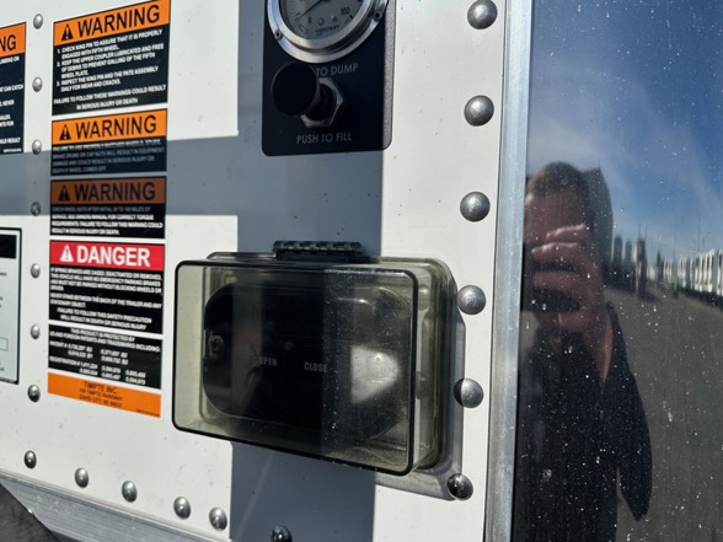 Trailer access panel and warning labels on a Timpte Super Hopper W trailer; visible text includes WARNING and DANGER labels, with an orange-dial gauge above and a black rectangular access door. The photo shows riveted metal paneling and a r