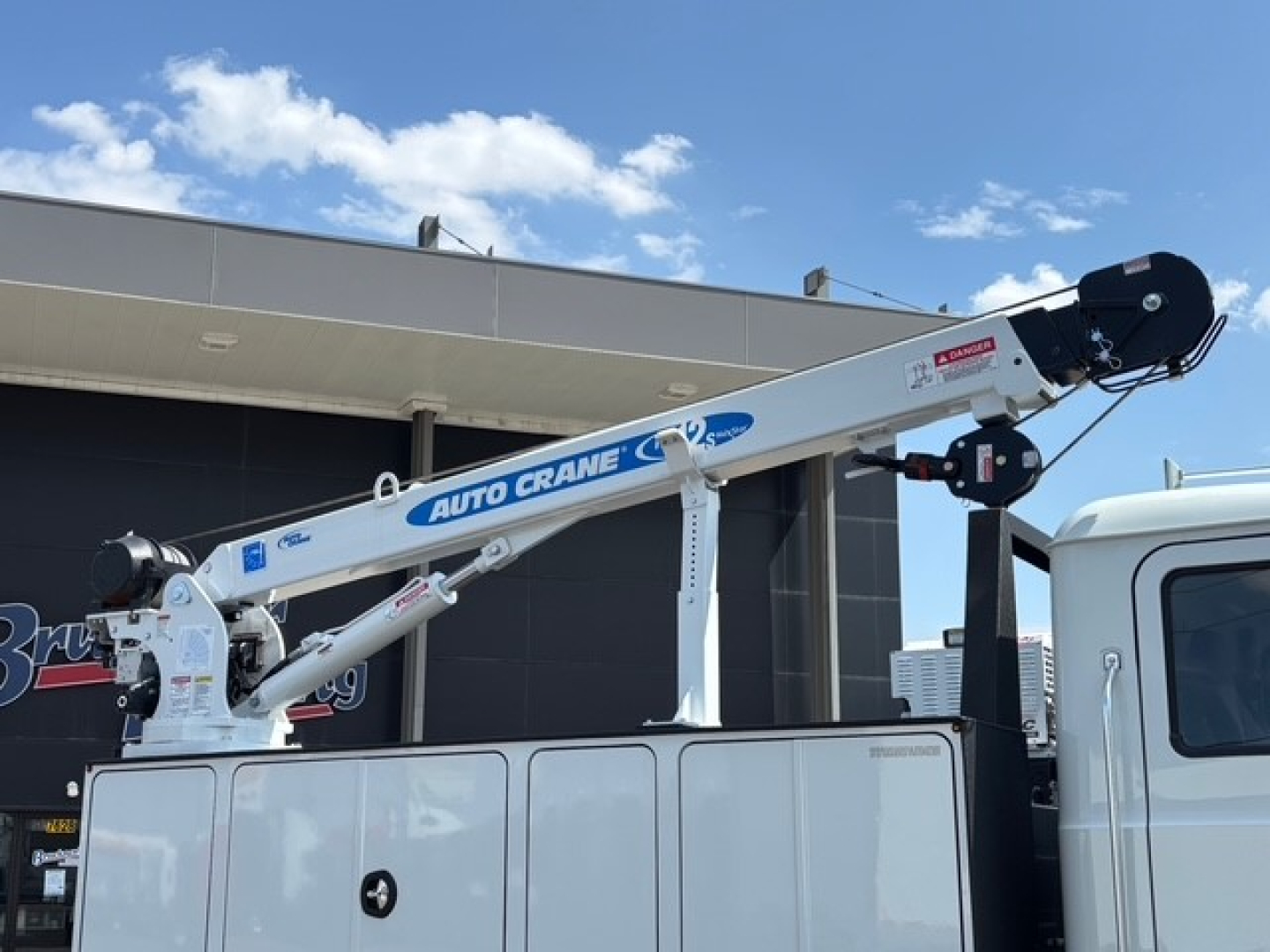 Service/utility truck with a white Auto Crane boom mounted on a Mack MD6, visible labels and hydraulic components, cab on the right, blue sky.